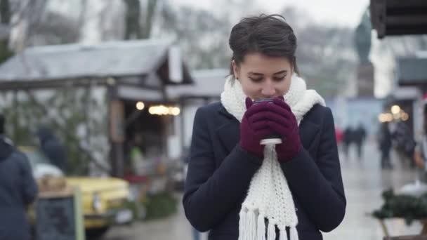 Portré bájos kaukázusi fiatal nő áll a téli utcában egy csésze kávé és mosolygós. Boldog lány fehér sállal és kabátban várja a barátját vagy férjét a szabadban. — Stock videók