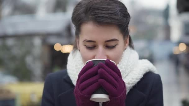 Close-up of pretty Caucasian woman drinking coffee and looking at camera. Beautiful girl in white winter scarf standing outdoors. Lifestyle, beauty, joy. — 비디오