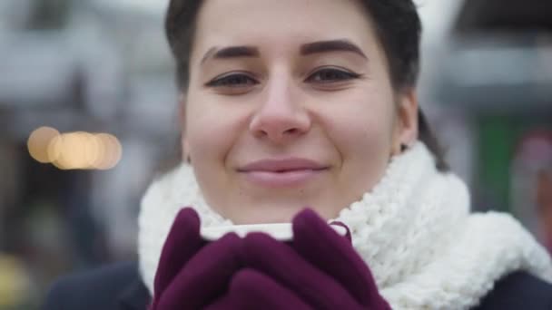 Extremo close-up de mulher morena caucasiana com olhos castanhos olhando para a câmera e degustação de café quente. Menina sorrindo no cachecol branco posando ao ar livre. Estilo de vida, felicidade, lazer . — Vídeo de Stock