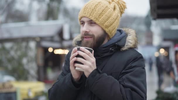 Egy fiatal, szakállas fehér férfi portréja vicces sárga kalapban és télikabátban, amint forró kávéval melegszik fel a szabadban. Fagyott jóképű fickó áll a téli vásáron és mosolyog. Szabadidő, életmód, boldogság. — Stock videók