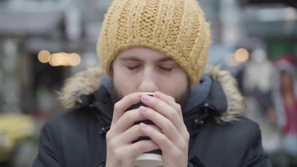 Κοντινό πλάνο πορτραίτο του παγωμένου νεαρού Καυκάσου που πίνει ζεστό καφέ στο δρόμο. Χαρούμενος τύπος με κίτρινο καπέλο και χειμωνιάτικο παλτό κοιτάζοντας την κάμερα. Αναψυχή, τρόπος ζωής. — Αρχείο Βίντεο