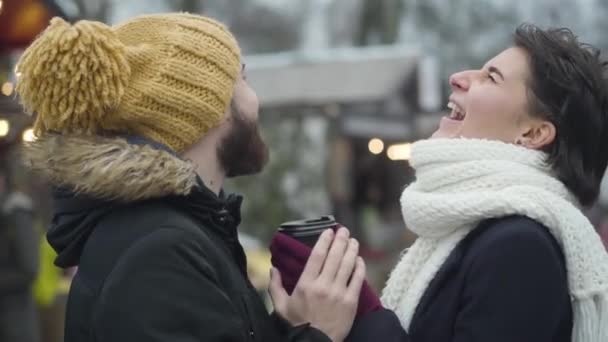 Vue de côté portrait de l'homme et de la femme caucasiens joyeux debout avec tasse de café sur la foire d'hiver et rire. Joyeux jeune famille se reposant ensemble à l'extérieur. Loisirs, mode de vie, détente . — Video