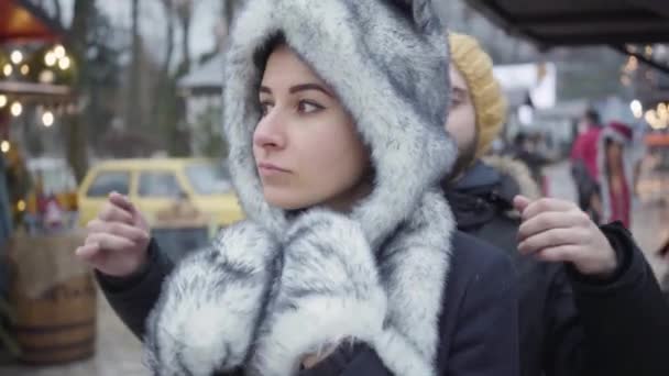 Retrato de la encantadora mujer caucásica en sombrero blanco y negro y guantes de pie al aire libre como hombre que viene de fondo y cerrando los ojos con las manos. Feliz familia joven divirtiéndose en feria de invierno . — Vídeos de Stock