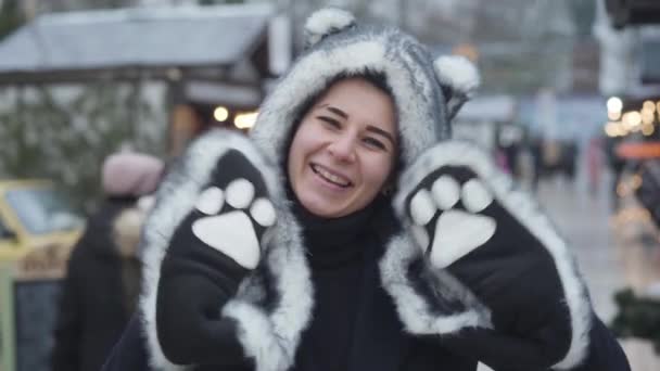 Porträtt av glad vit kvinna i varghatt och handskar ler och vinkar åt kameran. Glad ung flicka tillbringar helger på vintermarknad. Fritid, vila, livsstil. — Stockvideo