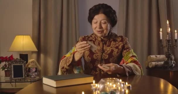 Madura mujer caucásica levantando jota de corazones, sonriendo y hablando. Feliz anciana adivina sentada en la mesa y leyendo el futuro. Sede del cine 4k ProRes . — Vídeos de Stock