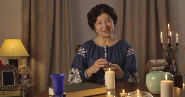 Портрет старої кавказької жінки в блакитній блузці, що сидить за столом зі свічками і картками. Посміхаючись, відьма кладе карти на стіл і розмовляє. Пророцтва, Августи, містики. Фільм 4k Prores Hq — стокове відео