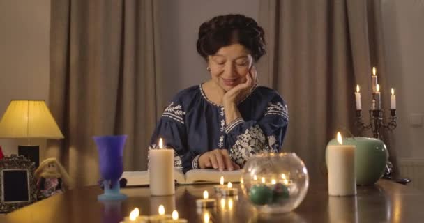 Retrato de una vieja morena caucásica sentada a la mesa con velas encendidas y leyendo. Vidente prediciendo el futuro con el uso del libro vintage. Sede del cine 4k ProRes . — Vídeo de stock