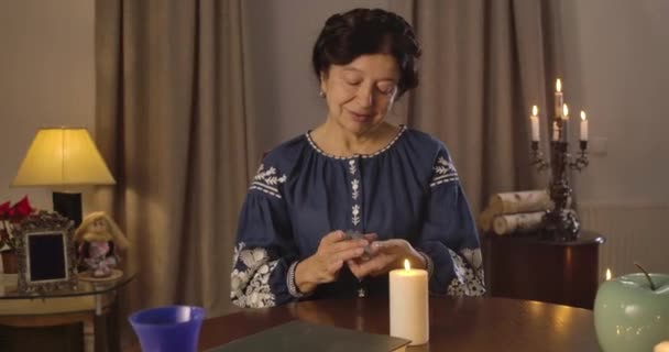 Camera beweegt van links naar rechts rond volwassen blanke vrouw schuifelen kaarten aan de tafel. Volwassen heks praat met kaarten, steekt kaars aan op de voorgrond. Bioscoop 4k Prores Hq. — Stockvideo