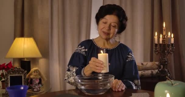 Зрелая белая женщина держит большую свечу и перемещает ее по тарелке с водой. Старшая волшебница, исполняющая ритуал предсказания судьбы. Магия, судьба, судьба. Штаб-квартира кинотеатра 4k ProRes . — стоковое видео