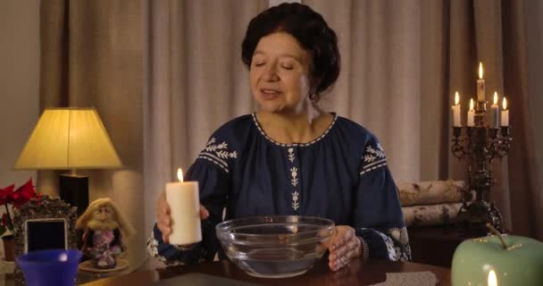 Old Caucasian brunette woman putting lighting candle on the table and moistening hands in water. Magician performing ritual to tell the destiny. Cinema 4k ProRes HQ. — 비디오
