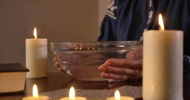 Zbliżenie żeńskich rąk kaukaskich kładących płytkę z wodą na stole, zapalających świecę i poruszających nią po cieczy. Dojrzała, nierozpoznawalna kobieta wykonująca rytuał. Kino 4k Prores Hq. — Wideo stockowe