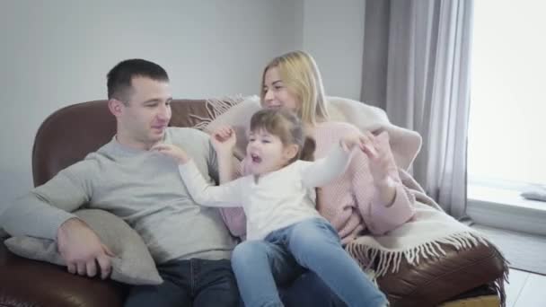 Retrato de menina branca bonito sentado em mãos mães, em seguida, abraçando o pai. Jovem família alegre desfrutando de tempo livre em casa juntos. Lazer, felicidade, paternidade . — Vídeo de Stock