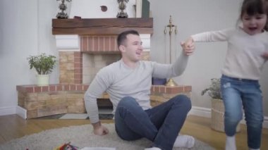 Neşeli küçük beyaz kız etrafta koşuşturup elini tutan, gülümseyen babasıyla sevgili kızını eline alan yetişkin bir adam. İçeride eğlenen mutlu ebeveyn ve çocuk portresi. Kamera yakınlaştırılıyor.