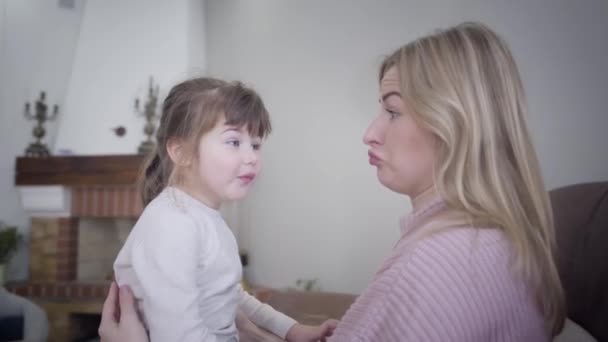 Retrato de mãe e filha caucasiana brincalhão rindo em casa. Mulher loira e pequena menina morena fazendo rostos e rindo como sentados na poltrona juntos. Divertimento, lazer, estilo de vida . — Vídeo de Stock