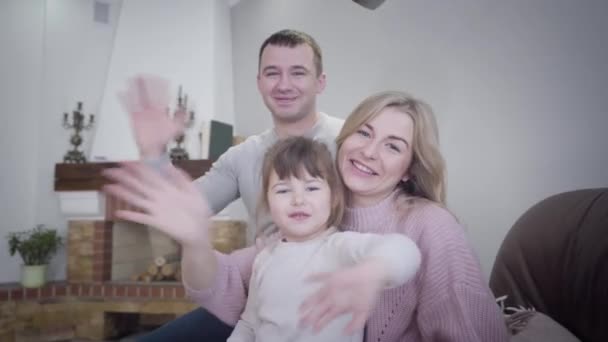 Portret van een gelukkige blanke familie die zwaait naar de camera. Glimlachende man, vrouw, en klein meisje zittend op een fauteuil binnen. Lifestyle, vreugde, geluk. — Stockvideo