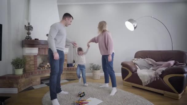 Joven esposo y esposa caucásicos tomados de las manos de sus hijas y criando hijos. Familia positiva juguetona disfrutando del tiempo juntos en el interior. Unidad, estilo de vida, felicidad . — Vídeo de stock