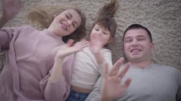 Beyaz bir ailenin yumuşak halıda uzanmış kameraya el sallamasının üst görüntüsü. Mutlu baba, anne ve kızın portresi. Yaşam tarzı, birlik, mutluluk. — Stok video