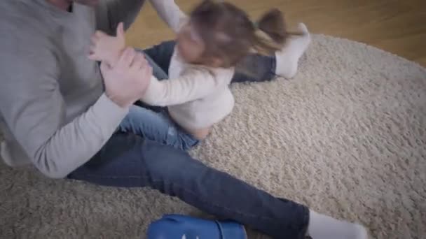 Ovanifrån av vacker brunett vit flicka leker med far hemma. Ung man har kul med dottern sittande på mjuk matta inomhus. Fritid, enhet, livsstil. — Stockvideo