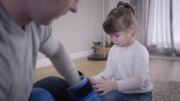 Niña caucásica concentrada poniendo guantes de boxeo en manos de padres irreconocibles. Bonita hija morena ayudando a su padre en interiores. Alegría, ocio, unidad . — Vídeos de Stock