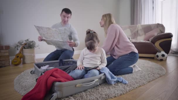 Retrato de una guapa morena caucásica jugando en un smartphone mientras sus padres se preparan para el viaje al fondo. Joven hombre y mujer examinando mapa y haciendo las maletas de viaje. Turismo, estilo de vida . — Vídeo de stock