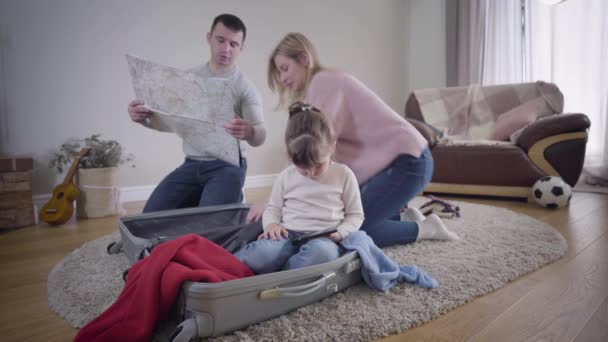 Jeune couple caucasien regardant la carte et discuter de futurs voyages comme leur jolie fille assise dans la valise et en utilisant un smartphone. Femme blonde souriante essayant beau châle. Tourisme, loisirs, unité . — Video