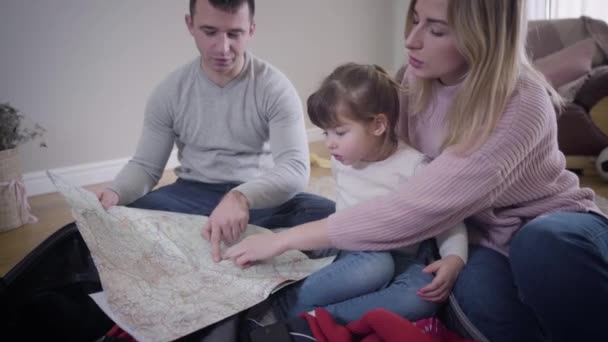 Porträtt av glada unga kaukasiska familjen gör rutten för framtida resa. Leende ung man, kvinna och liten söt flicka sitter hemma och tittar på kartan prata. Turism, resor. — Stockvideo