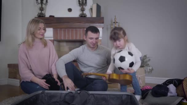 Serieuze kleine blanke meisje zet ukelele en bal in koffer en helpt moeder om de reistas te sluiten. Mooie dochter bereidt spullen voor om met ouders te reizen. Lifestyle, kindertijd, toerisme. — Stockvideo