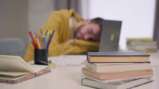 Młody, biały student budzi się i zaczyna pisać na klawiaturze laptopa. Zmęczony przystojniak przepracowany. Skup się na zmianach od książki na pierwszym planie do twarzy człowieka. — Wideo stockowe