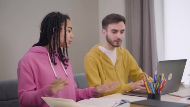Schattig Afrikaans Amerikaans meisje met afro vlechtjes die naast een knappe blanke jongen zitten met een laptop. Jonge groepsgenoten discussiëren over onderwijs. Intelligentie, levensstijl, studeren. — Stockvideo