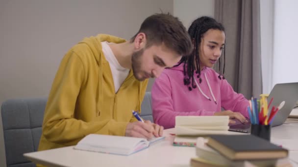 Portret van vrolijke studenten die aan tafel zitten en studeren. Afro-Amerikaans meisje praat met blanke jongen en glimlacht. Een man schrijft, een vrouw gebruikt een laptop. Intelligentie, levensstijl, onderwijs. — Stockvideo