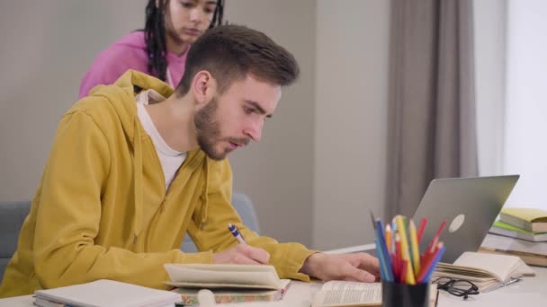 Atractiva chica afroamericana trayendo una taza de té o café para el joven chico caucásico sentado en la mesa leyendo y escribiendo. Estudiante ayudándose mutuamente a estudiar. Educación, estilo de vida . — Vídeos de Stock