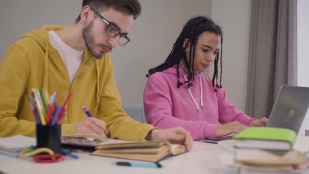 Стресс афро-американская девушка закрывает ноутбук и скрещивает руки, как ее мужчина европеец в очках закатывает глаза и пишет в книге. Образование, трудности, учеба . — стоковое видео