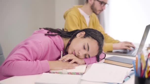 Close-up retrato de jovem bela menina afro-americana dormindo na mesa como menino caucasiano inteligente em óculos digitando no laptop no fundo. Estudantes que trabalham demais na universidade . — Vídeo de Stock