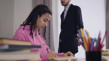 Side view portrait of attractive African American girl writing down as young Caucasian teacher in suit walking at the background and dictating. College, university, studying.