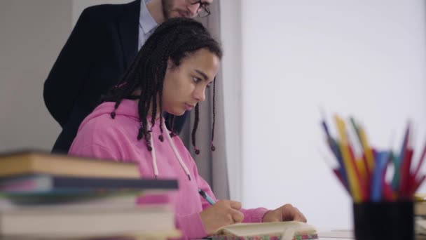 Joven chica afroamericana sentada en la mesa y anotando la conferencia de profesores. Hombre caucásico maestro en traje caminando en el fondo y mirando en los estudiantes notas sobre su hombro . — Vídeos de Stock