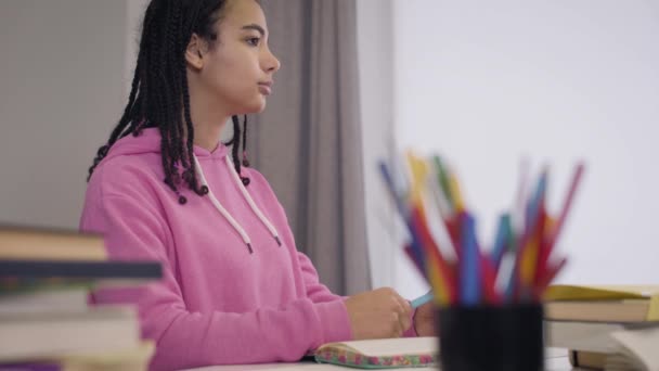 Side view of beautiful young African American girl sitting at the table and listening carefully to professor. Portrait of pretty female student writing down notes in workbook. Intelligence, education. — Stock Video