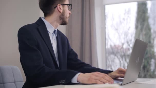 Portrait de jeune homme blanc beau regardant par la fenêtre et tapant sur le clavier de l'ordinateur portable. Homme d'affaires confiant en costume et lunettes de travail en ligne. Mode de vie, intelligence, richesse . — Video