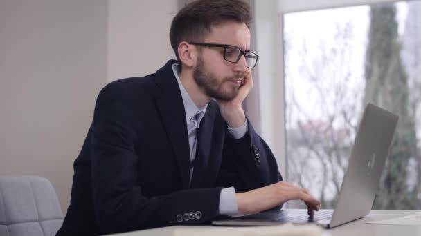 Jeune homme d'affaires caucasien fatigué tenant la tête avec la main et tapant sur le clavier de l'ordinateur portable. Il s'ennuyait à enlever les lunettes et à les mettre sur la table. Entreprise, travail, freelance . — Video