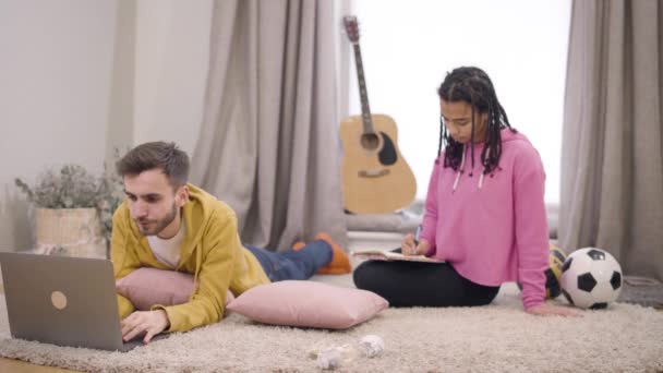 Portrait of cheerful young multiethnic couple resting at home on weekends. Handsome Caucasian boy using laptop and African American girl writing at the background. Happiness, lifestyle, leisure. — Stock Video