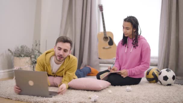 Handsome Caucasian boy showing something interesting on laptop screen to pretty African American girl writing at the background. Smiling man and woman resting indoors. Happiness, leisure, lifestyle. — Stok video