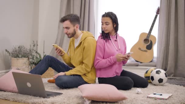 Young multiethnic couple sitting back to back on the floor and using smartphones. Girlfriend spying boyfriend over shoulder. African American woman and Caucasian man with Internet addiction. — 비디오