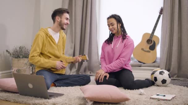 Jeune garçon caucasien souriant chantant sérénade et jouant ukulélé pour sa petite amie afro-américaine. Couple amoureux passer du temps libre ensemble à l'intérieur. Amour, bonheur, romance . — Video