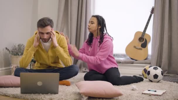 Retrato do jovem caucasiano estressado sentado na frente do laptop segurando a cabeça com as mãos e ouvindo a mulher afro-americana falando emocionalmente. Garoto exausto passando tempo com a namorada . — Vídeo de Stock