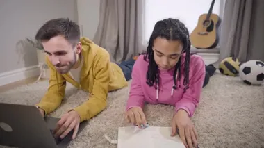 Concentrated African American girl writing and putting hand on floor as Caucasian boy touching her palm and smiling. Young students in love doing homework together indoors. Lifestyle, happiness.
