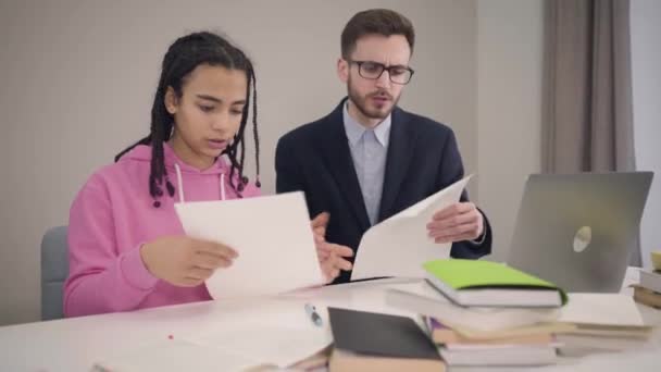 Camera approaching slowly to African American girl and Caucasian boy looking through documents. Young students helping papers and talking with each other. Lifestyle, intelligence, education. — Stockvideo
