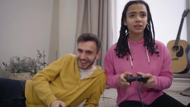 Bello ragazzo caucasico che perde nel videogioco mentre la sua ragazza afroamericana vince e sorride. Due giovani allegri che giocano insieme in casa. Hobby, stile di vita, gioia . — Video Stock