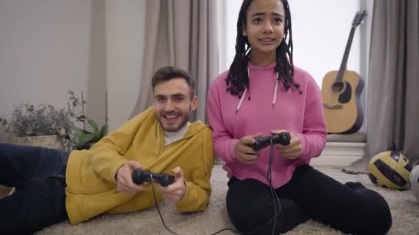 Camera moving up, cute African American girl losing in video game, Caucasian winner smiling and hugging his upset girlfriend. Lifestyle, leisure, gaming. — Stockvideo