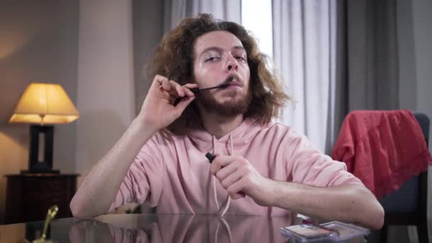 Πορτρέτο του συγκεντρωμένου δυαδικού φύλου Καυκάσιος που εφαρμόζει μάσκαρα στα μουστάκια και χαμογελά στην κάμερα. Άνθρωπος με μακιγιάζ στη μία πλευρά του προσώπου. Φύλο, ταυτότητα, ουδετερότητα. — Αρχείο Βίντεο