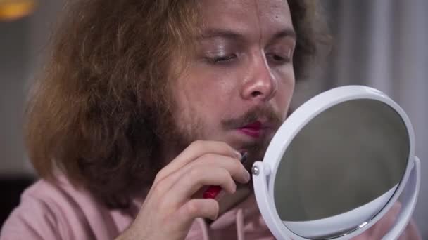 Close-up portrait of young Caucasian man with face divided into two parts applying red lipstick. Expression of intersex person self perception. — ストック動画