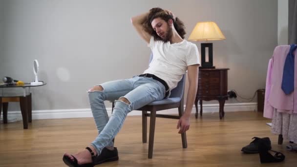 Retrato de tiro médio de homem branco bonito jovem sentado na cadeira tocando o cabelo e olhando para a câmera. Intersex pessoa vestindo uma bota broggi masculino e um sapato de salto alto feminino . — Vídeo de Stock